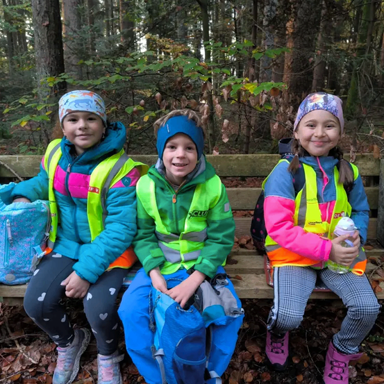 Drei Kinder sitzen auf einer Bank im Wald. Sie tragen alle neongrüne Westen. Das Mädchen auf der linken Seite hat einen Rucksack und lächelt. Der Junge in der Mitte hat eine blaue Mütze und schaut zur Seite. Das Mädchen auf der rechten Seite hat eine Wasserflasche in der Hand.