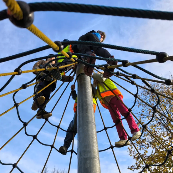 Vier Personen klettern eine Seilnetzstruktur. Zwei tragen Sicherheitswesten. Die Szene ist draußen mit blauem Himmel und Bäumen.
