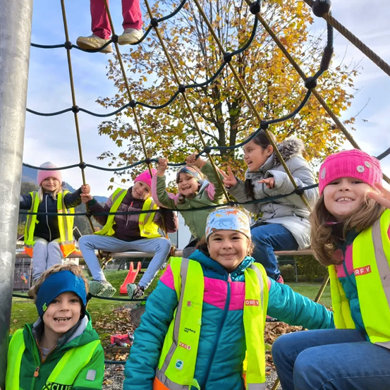 Kinder in Warnwesten spielen auf einem Spielplatz, eines klettert auf einer Seilleiter und andere lächeln und winken. Dahinter sitzt ein Mädchen auf einer Bank und ein Baum mit gelben Blättern.