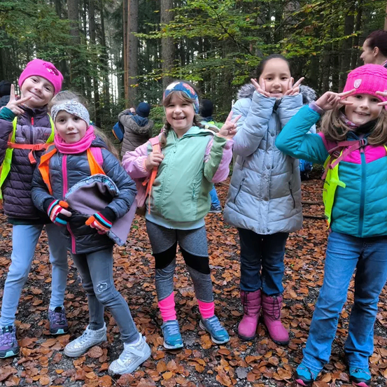 Fünf junge Mädchen in bunter Winterkleidung posieren für ein Foto im Wald. Sie machen alle Friedenszeichen und lächeln. Der Boden ist mit trockenen Blättern bedeckt.