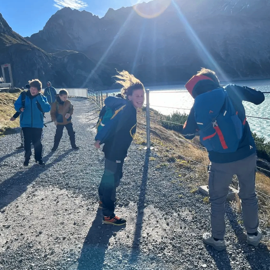 Eine Gruppe von Wanderern mit Rucksäcken geht auf einem Schotterweg am See entlang, mit Bergen im Hintergrund.