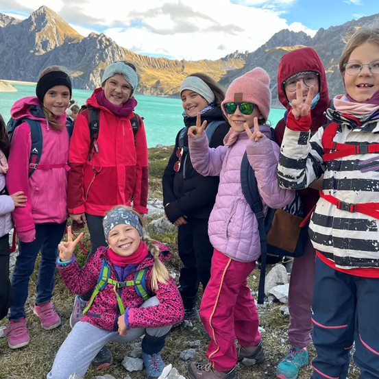 Eine Gruppe von Kindern in Winterkleidung posiert für ein Foto am See mit Bergen im Hintergrund. Einige tragen Mützen und Brillen, alle haben Rucksäcke.