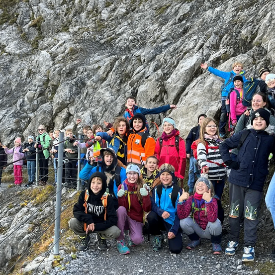 Eine Gruppe von Kindern und Erwachsenen posiert für ein Foto vor einem felsigen Berg. Sie tragen Winterkleidung und einige haben Rucksäcke. Sie lächeln und machen Gesten.