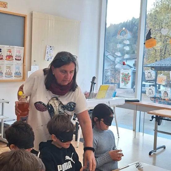 Eine Frau in einem Klassenzimmer schenkt getränke für verblindete Kinder an einem Tisch ein. Sie halten Gläser und es gibt eine Tafel und ein Fenster mit Dekorationen.