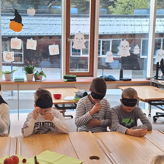 Mehrere Kinder sind mit verbundenen Augen und trinken aus Gläsern an einem Tisch. Dahinter befindet sich ein Fenster mit Halloween-Dekorationen und Pflanzen.
