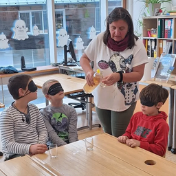 Eine Frau gießt Flüssigkeit in ein Glas, während drei verbundene Kinder vor ihr am Tisch sitzen, möglicherweise an einer sensorischen Aktivität teilnehmend.