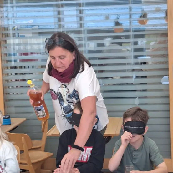 Eine Frau mit Brille gießt Saft in einen Becher, während zwei vermummte Kinder ihn auf einem Tisch probieren.