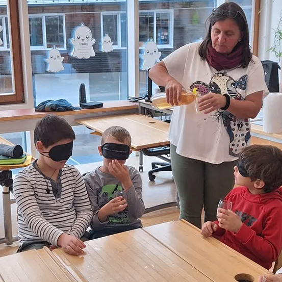 Eine Frau gießt ein Getränk in ein Glas, während drei mit verbundenen Augen wartende Kinder sich möglicherweise an einer sensorischen Aktivität beteiligen.