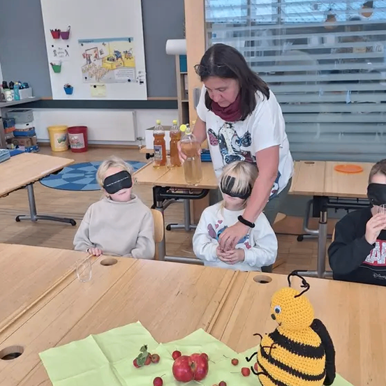 Eine Frau leitet blindgebundene Kinder in einem Klassenzimmer, wobei sie einen Tisch mit einer gehäkelten Biene und Früchten für eine sensorische Aktivität benutzt.