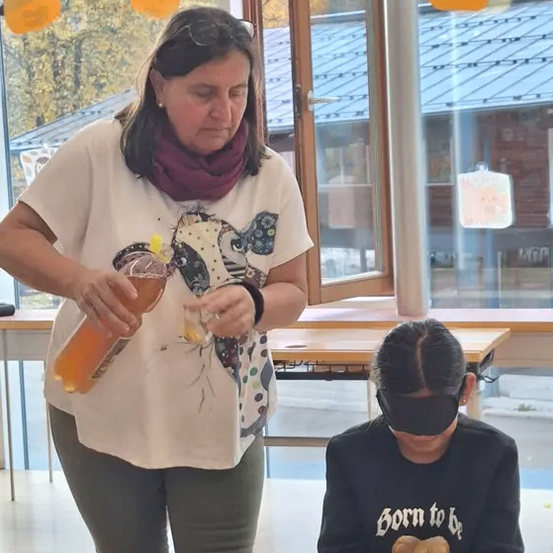 Eine Frau mit einer Augenbinde sitzt an einem Tisch, während eine andere Frau Flüssigkeit in ein Glas gießt. Die Szene spielt sich drinnen ab, mit Bäumen, die durch ein Fenster sichtbar sind.