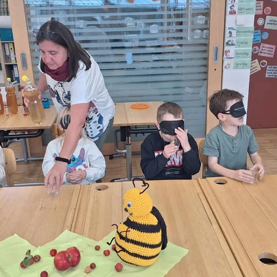 Drei Kinder und eine erwachsene Frau sind in einem Klassenzimmer, mit verbundenen Augen, trinken aus Gläsern. Der Tisch hat eine gehäkelt Biene und einige rote Früchte.