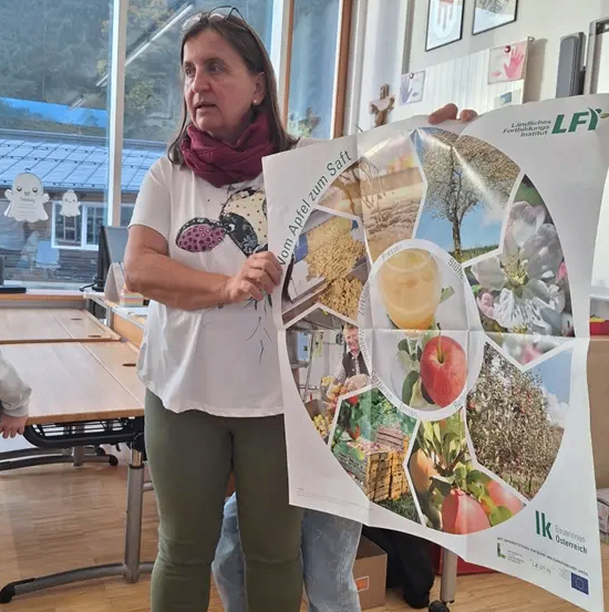 Eine Frau in einem weißen T-Shirt und grüner Hose hält ein großes Poster mit einer Collage von Äpfeln und anderen Früchten. Hinter ihr ist ein Klassenzimmer mit Tischen und einem Fenster zu sehen.