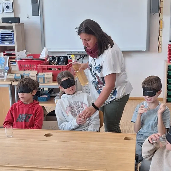 Eine Frau gießt eine Flüssigkeit in Gläser, die von verbundenen Kindern gehalten werden, die an einem Tisch sitzen. Sie befinden sich in einem Klassenzimmer mit einer Tafel und Regalen im Hintergrund.