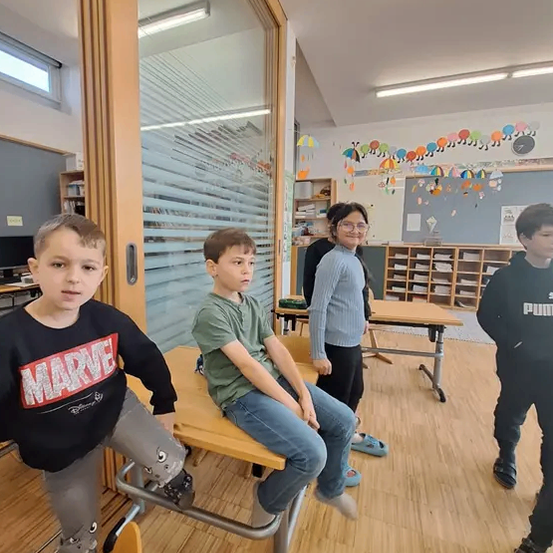 Eine Gruppe von Kindern steht in einem Klassenzimmer. Ein Junge mit einem Marvel-T-Shirt steht auf einem Schreibtisch, und ein anderer Junge in einem grünen Shirt sitzt auf dem Schreibtisch. Dahinter steht ein Mädchen mit Brille.