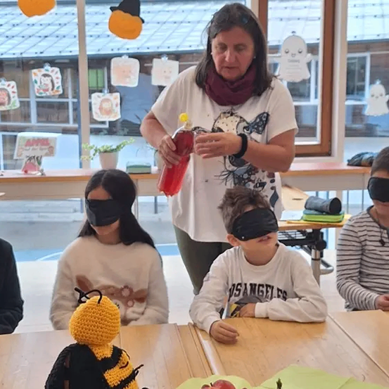 Eine Frau füllt Saft in eine Flasche, während ein verblindeter Junge und ein Mädchen mit einer gehäkelt Biene, Äpfeln und einer Kürbis am Tisch sitzen.