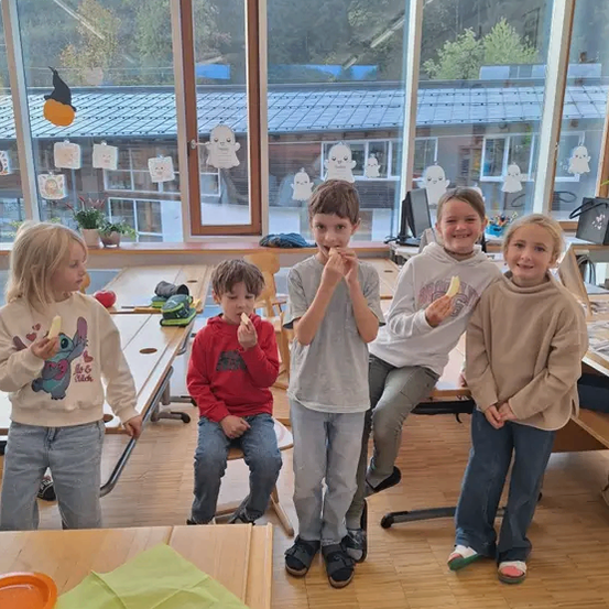 Fünf Kinder, in lässiger Kleidung gekleidet, stehen und sitzen in einem Klassenzimmer, jedes isst einen Apfel. Dahinter befindet sich ein großes Fenster mit Blick auf Bäume und Gebäude.