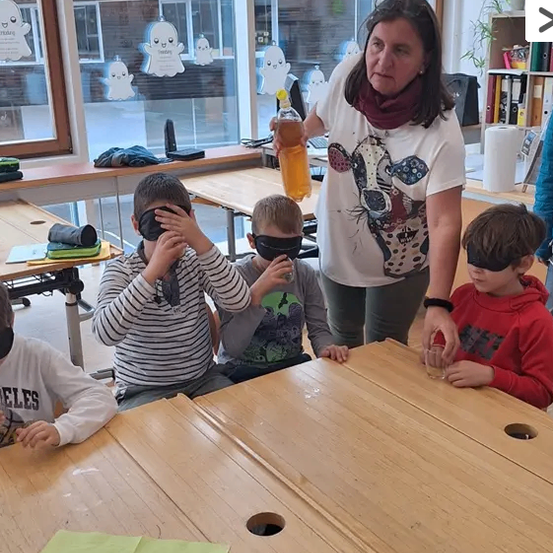 Eine Frau hält eine Flasche und vier verbundene Kinder sitzen an einem Tisch in einem Klassenzimmer.