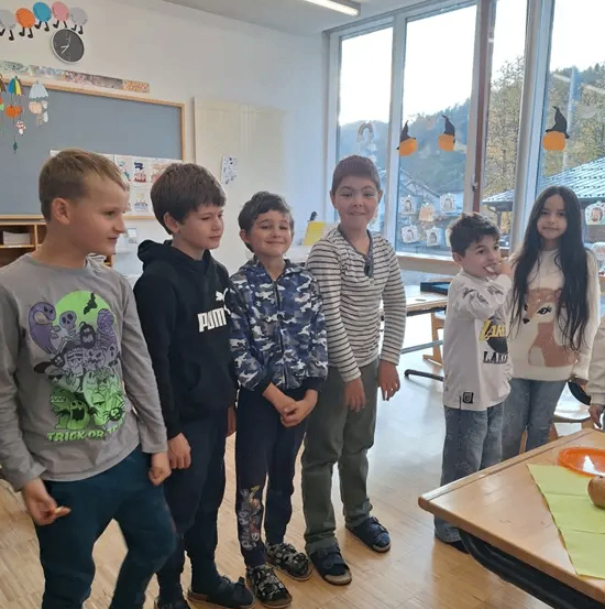 Sechs Kinder stehen in einem Klassenzimmer und lächeln. Sie tragen Freizeitkleidung. Hinter ihnen ist eine Tafel mit Dekorationen und einer Uhr zu sehen. Große Fenster lassen viel Licht herein.