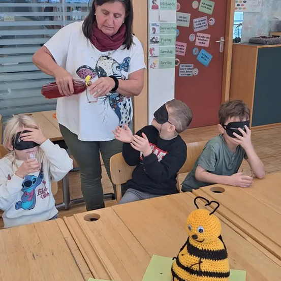 Eine Frau in einem weißen T-Shirt schenkt Kindern, die Augenbinden tragen, Saft ein, die an einem Tisch mit einer gehäkelt Biene und Äpfeln sitzen.