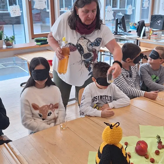 Eine erwachsene Frau unterrichtet drei verblindete Kinder in einem Klassenzimmer. Sie gießt Saft in ein Glas. Eine gehäkelter Biene und einige Früchte liegen auf dem Tisch.