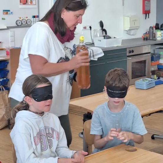 Eine Frau gießt eine Flüssigkeit aus einer Flasche in Gläser, die von zwei verbundenen Kindern gehalten werden, die an einem Tisch in einem Klassenzimmer sitzen.