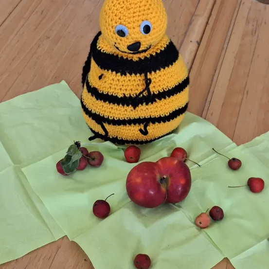 Eine gehäkelte Biene sitzt auf grünem Papier mit roten Kirschen und Äpfeln auf einem Holztisch.
