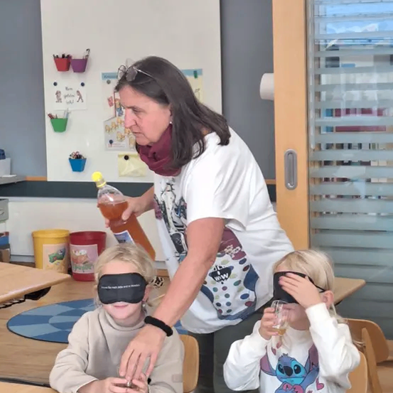Eine Frau gießt Saft in Gläser für zwei verbundene Kinder, die an einem Tisch in einem Klassenzimmer sitzen.