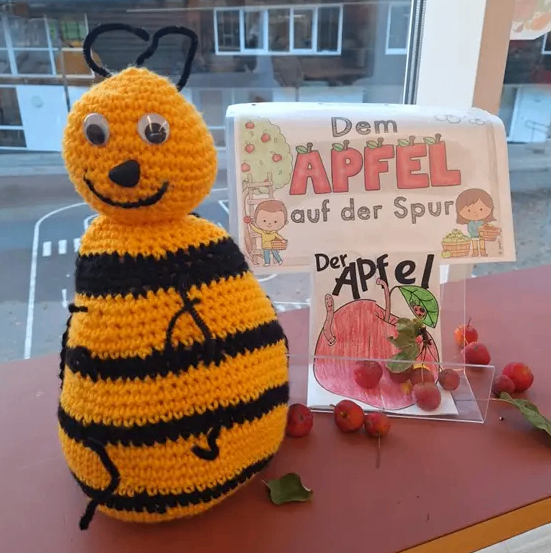 Ein gehäkeltes Bienchen sitzt auf einem Tisch neben einem Buch mit dem Titel 'Dem Apfel auf der Spur.' Das Buch wird von kleinen Äpfeln und Blättern begleitet. Hinter dem Plüsch reflektiert ein Fenster ein Gebäude mit Dach.