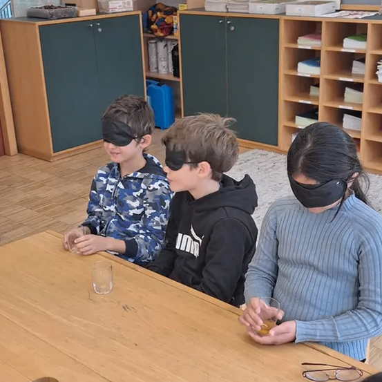 Drei verschleierte Kinder sitzen an einem Holztisch, jeder hält ein Glas. Sie nehmen an einer sensorischen Aktivität in einem Klassenzimmer teil. Hinter ihnen stehen Schränke mit Büchern und Aufbewahrungsboxen.