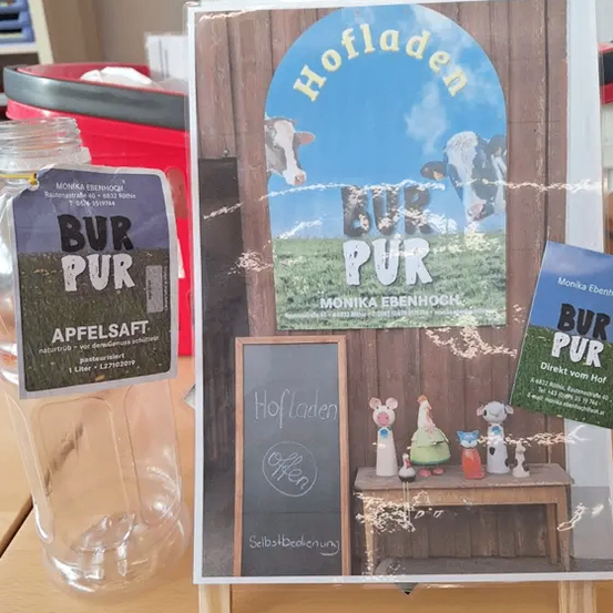 Auf einem Tisch steht ein Buch mit dem Titel 'Hofladden', eine Tafel mit dem Wort 'Open' und eine Glasflasche mit einem Etikett, das 'BUR PUR Apfelsaft' liest. Dahinter befindet sich ein Poster mit einer Kuh auf einem Feld.