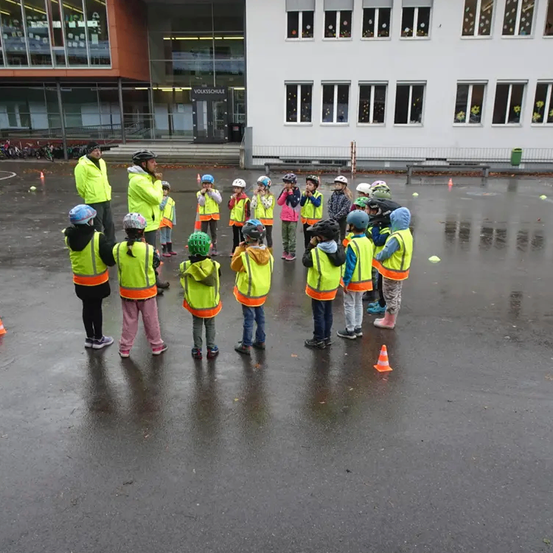 Mehrere Kinder in gelben Westen sind um einen Mann vor einem Gebäude versammelt. Sie tragen alle Helme und scheinen Anweisungen zu lauschen.