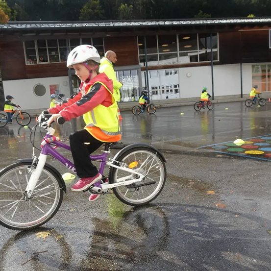 Ein junges Mädchen fährt ein lila Fahrrad auf einem nassen Gelände. Sie trägt eine gelbe Weste und einen Helm. In der Nähe fahren Kinder im Kreis auf Fahrrädern. Ein Mann steht hinter dem Mädchen.