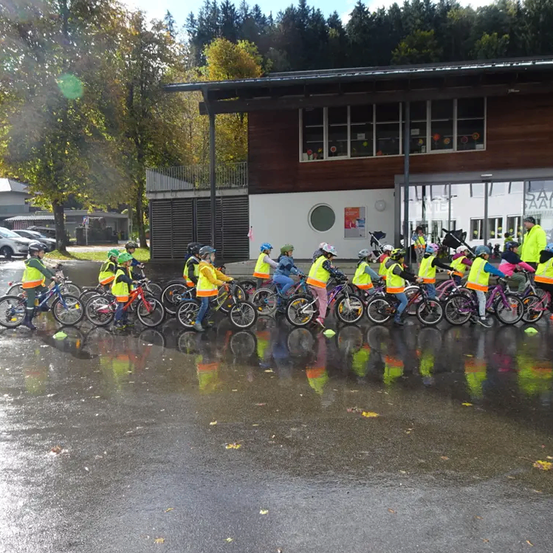 Eine Gruppe von Kindern in Neonwesten fährt in einer Reihe vor einem Gebäude mit Bäumen im Hintergrund auf Fahrrädern.