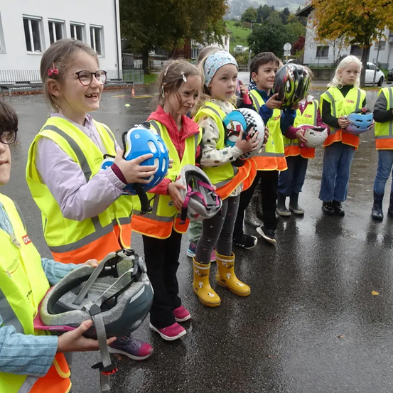 Eine Gruppe von Kindern in gelben Sicherheitswesten, die Helme halten, steht draußen im Regen und lächelt und posiert für ein Foto. Hinter ihnen steht ein Gebäude mit Fenstern.