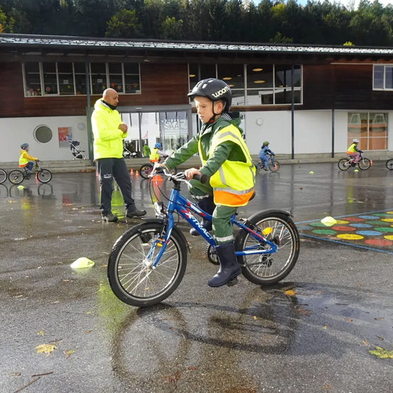 Ein junger Junge in einer gelb-grünen Jacke fährt ein blaues Fahrrad auf einer nassen Straße. Ein Mann in einer gelben Jacke beobachtet.