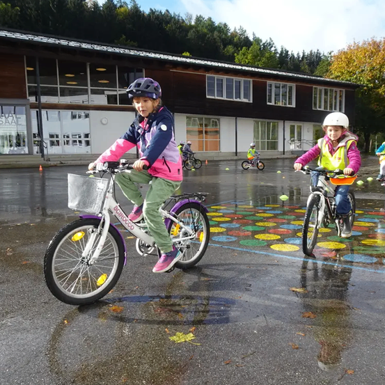 Zwei Kinder fahren mit ihren Fahrrädern auf einem nassen Parkplatz. Hinter ihnen befindet sich ein Gebäude mit Glasfenstern.