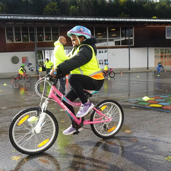 Ein junges Mädchen fährt auf einer nassen Straße ein pinkes Fahrrad und trägt eine gelbe Weste und einen Helm. Hinter ihr hilft ein Mann. In der Nähe fahren andere Kinder Fahrräder, und ein Gebäude mit Glasfenstern ist im Hintergrund.