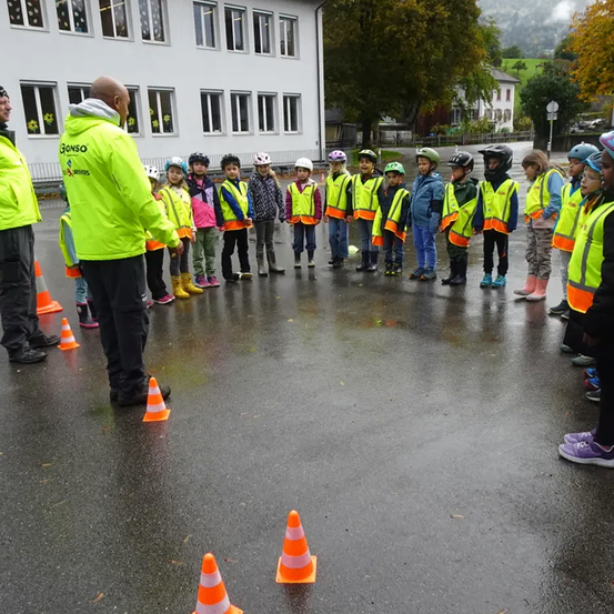 Eine Gruppe junger Kinder in reflektierenden Westen und Helmen steht in einem nassen Bereich, geführt von einem Mann in einer neongrünen Jacke. Der Bereich ist von Verkehrskegeln und einem Gebäude umgeben.