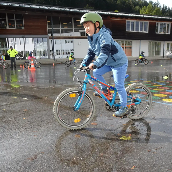 Ein junger Junge fährt auf einem blauen Fahrrad in einem nassen Parkplatz mit anderen Kindern. Er trägt einen grünen Helm und blaue Kleidung. Im Hintergrund befinden sich Gebäude und Bäume.