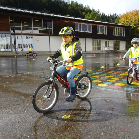 Zwei Kinder in Sicherheitswesten fahren auf einem nassen Parkplatz mit bunten Kreisen auf dem Boden mit Fahrrädern. Ein Gebäude mit Glaswänden steht dahinter.
