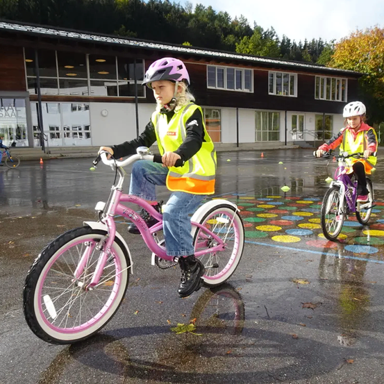 Zwei junge Mädchen fahren auf einer nassen Straße mit ihren Fahrrädern. Sie tragen Sicherheitswesten und Helme. Hinter ihnen ist ein Gebäude mit Glasfenstern zu sehen.