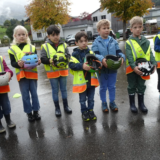 Eine Gruppe von Kindern in Sicherheitswesten hält Helme in einem nassen Parkplatz mit Häusern und Bäumen im Hintergrund.