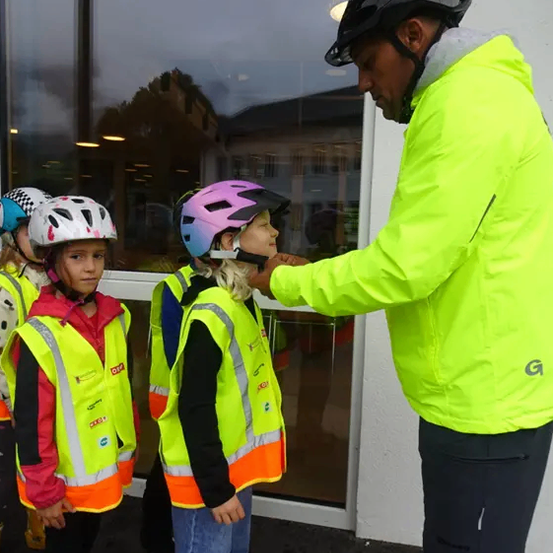 Ein Mann justiert den Helm eines Mädchens, während eine Gruppe von Kindern vor einer Glastür steht.