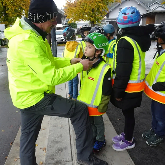 Ein Mann in einer neongelben Jacke hilft einem Kind mit einem grünen Helm, es anzupassen. Dahinter stehen mehrere andere Kinder in neonfarbenen Westen und Helmen.