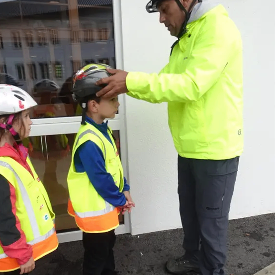 Ein Mann hilft einem Jungen, seinen Helm vor einer Glastür nachzujustieren, wobei ein Mädchen in einer reflektierenden Weste in der Nähe steht.