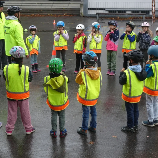 Eine Gruppe von Kindern in reflektierenden Westen und Helmen steht im Kreis mit zwei Erwachsenen, möglicherweise lernen sie Fahrrad fahren.
