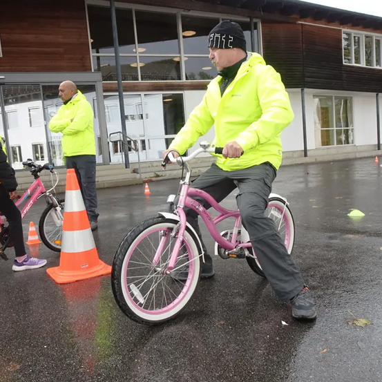 Ein Mann in einer neongelben Jacke fährt ein pinkes Fahrrad, mit zwei anderen Personen in neongelben Jacken und Verkehrskegeln im Hintergrund.