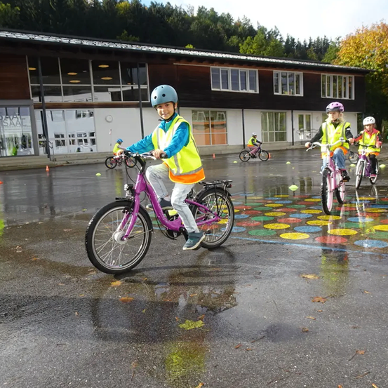 Kinder, die Helme tragen, fahren auf einer nassen Straße mit einem mehrfarbigen Kreismuster vor einem Gebäude mit Fahrrädern.