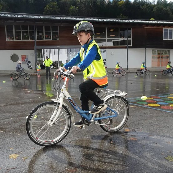Ein junger Junge fährt ein Fahrrad auf einer nassen Straße, trägt einen Helm und eine Warnweste. Hinter ihm sind andere Kinder, die Fahrräder fahren. Die Szene spielt sich in einer Schule ab, mit einem Gebäude im Hintergrund.