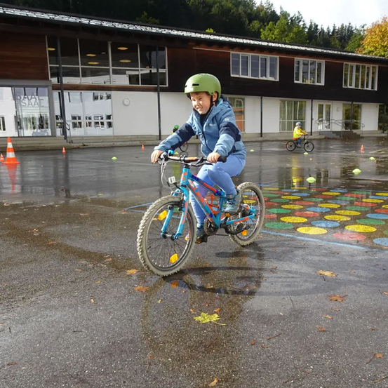 Ein Kind mit Helm fährt auf einer nassen Straße mit dem Fahrrad, im Hintergrund steht ein Gebäude und es sind Bäume zu sehen.